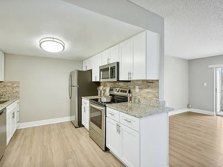Modern kitchen with granite countertops, stainless steel appliances, and ample white cabinetry, adjacent to a bright living area with sleek hardwood floors and balcony access.