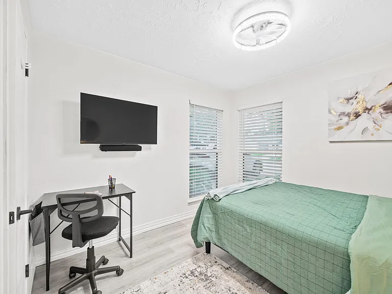 Furnished bedroom with a work desk, wall-mounted TV, and two windows with blinds for natural light and privacy.