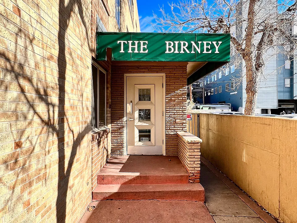 Inviting entrance of The Birney apartment building with a charming brick facade and green awning under a clear blue sky.