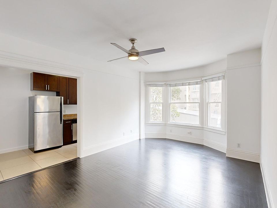 Spacious empty room with hardwood floors, ceiling fan, and large bay windows. Open kitchen with stainless steel fridge and cabinets.