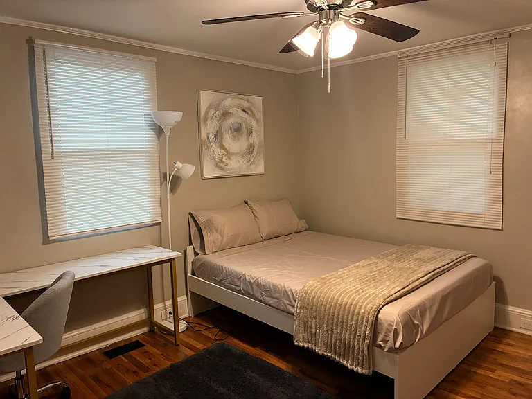 Furnished bedroom with a full bed, desk workspace, ceiling fan, and two windows with blinds for light and privacy.