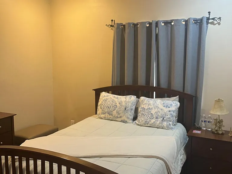 Furnished bedroom with a queen bed, nightstand, dresser, ceiling fan, and window with full curtains for privacy.