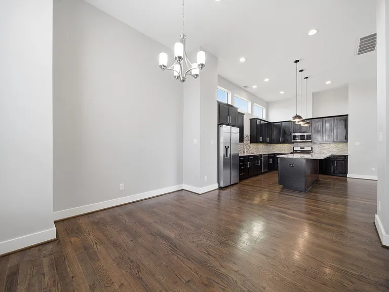 Stunning modern kitchen and dining area with high ceilings, rich dark wood flooring, and bold black cabinetry. A spacious island with pendant lighting anchors the space, complemented by stainless steel appliances and large windows.