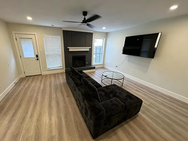 Open living room with wood-style flooring, ceiling fan, fireplace, and wall-mounted TV, plus nearby exterior access.