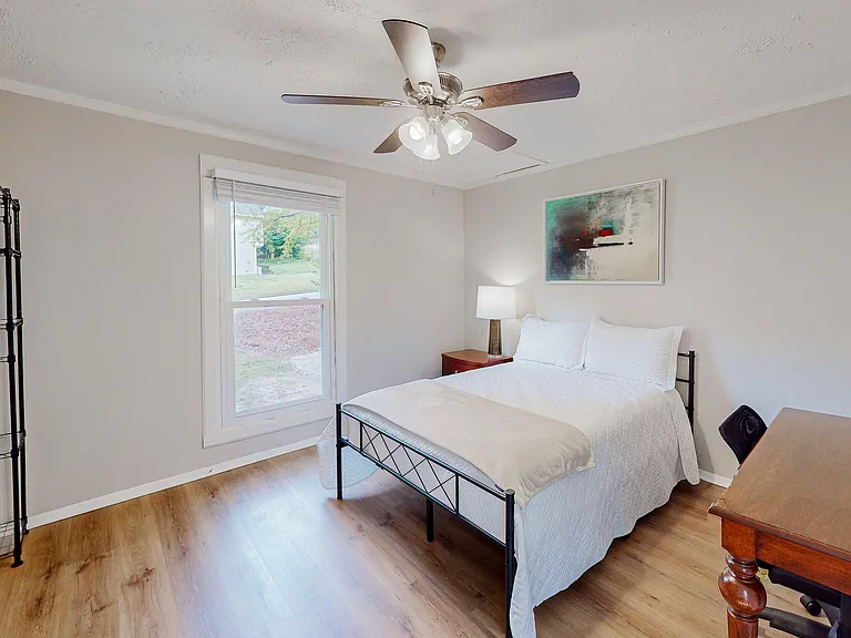 Furnished bedroom with a full bed, ceiling fan, large window, and workspace, plus open shelving for storage.