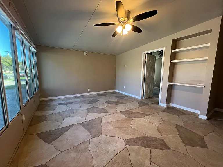 Entering the front door to the living room with plenty of windows to allow in light with full blinds for privacy. Wall is ready to mount a television. Small desk area with shelving.