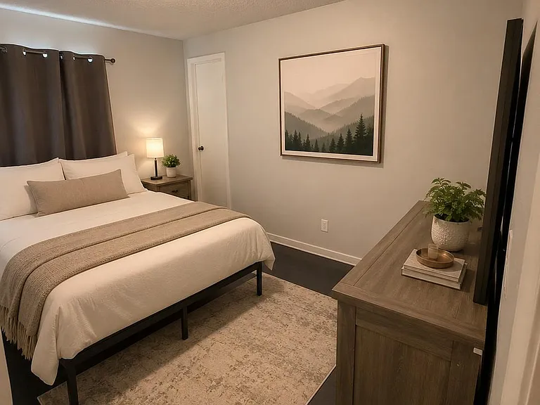 Furnished bedroom with a queen bed, nightstand lighting, dresser/TV setup, and a large area rug for added comfort.