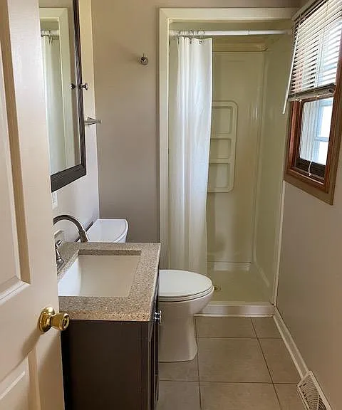 Bathroom with a window for natural light, medicine cabinet and shelf above toilet. Shower in background. Tile Floor.
