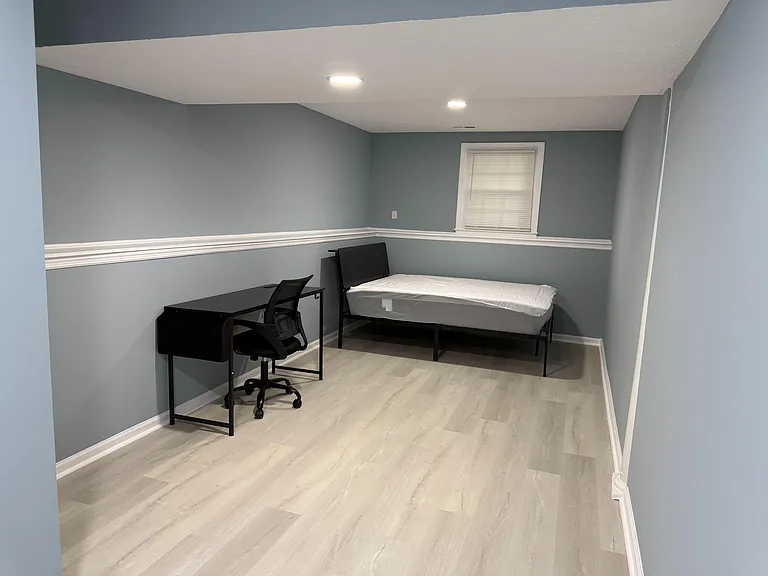 Furnished bedroom with vinyl plank flooring, neutral paint, overhead lighting, and a workstation plus window with blinds.
