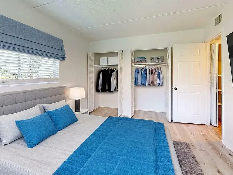 Bright and airy bedroom featuring a plush gray bed with blue accents, dual closets with contemporary wardrobe, and a wall-mounted flat-screen TV.