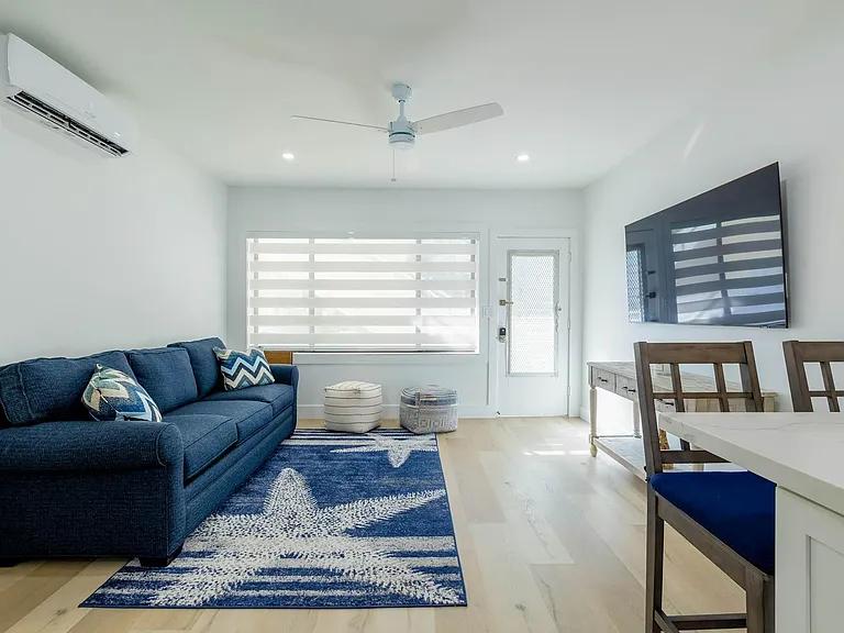 Open living area with plush seating, wall-mounted TV, ceiling fan, and large window with privacy shades.