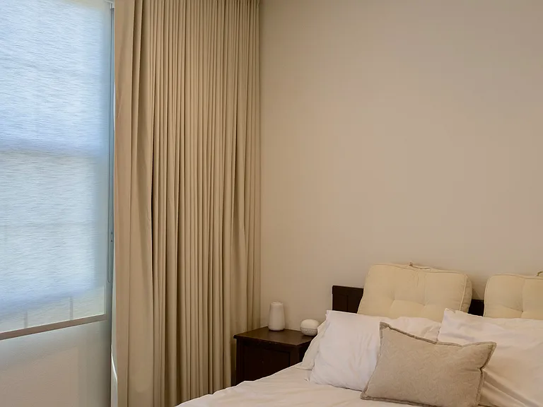 Primary Bedroom: Corner view highlighting the window setup with included full-length blackout drapery, positioned right beside the bedside nightstand.