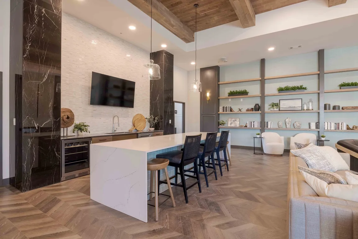The clubhouse demonstration kitchen features a quartz waterfall island with seating for four, a built in wine refrigerator, and open shelving for hosting or everyday use.