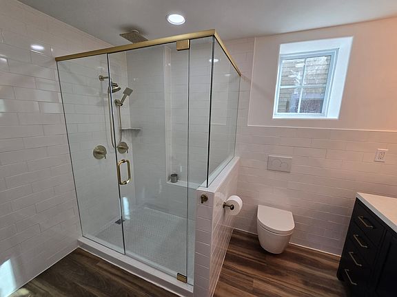 Modern bathroom with glass-enclosed shower featuring double showerheads, gold fixtures, white subway tiles, wall-mounted toilet and window lettings tons of natural light in.