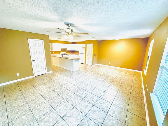 View from the entrance into a tiled living space with beige walls, ceiling fan, and open kitchen featuring white cabinets, a pantry, closet, appliances. Clean, modern layout with two windows allowing natural light.
