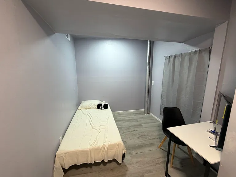 Compact bedroom with a twin bed, durable flooring, and a work desk setup, plus window coverage for privacy.