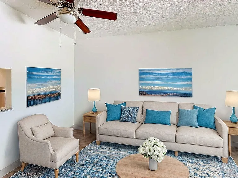 Charming living room with a cozy beige sofa, vibrant blue cushions, elegant armchair, and modern ceiling fan. Features a stylish open concept design with a glimpse into a contemporary kitchen.