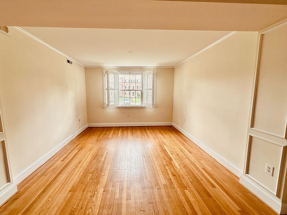 Bright and airy room with hardwood floors, crown molding, and neutral walls. A large window with plantation shutters fills the space with light and offers charming views, making it perfect for living, dining, or versatile use.