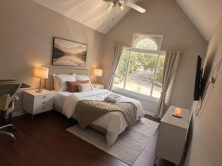 Vaulted bedroom features a queen bed, nightstands with lighting, a work desk, and a large arched window with curtains.