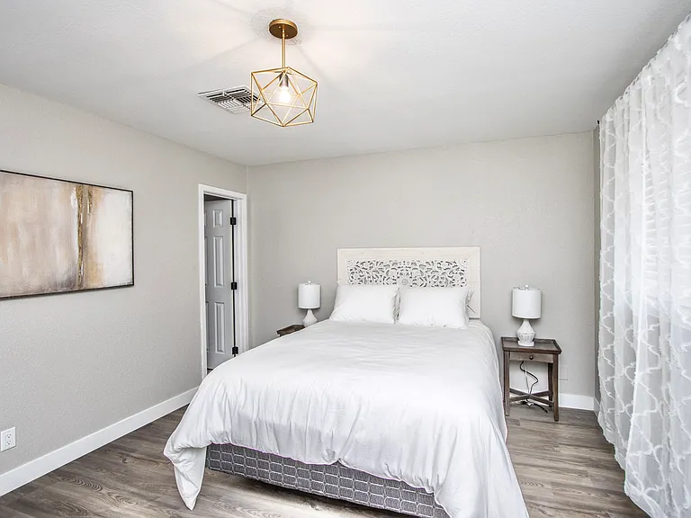 Serene primary bedroom at Casa Del Sol, Tempe rental home. Features a comfortable bed with a carved wood headboard, a large rustic full-length mirror, a blue accent chair, and modern geometric lighting. A peaceful Tempe, AZ corporate retreat.