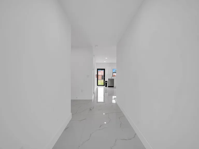 Long hallway with polished tile flooring and clean white walls, leading to a rear door and adjacent kitchen area.
