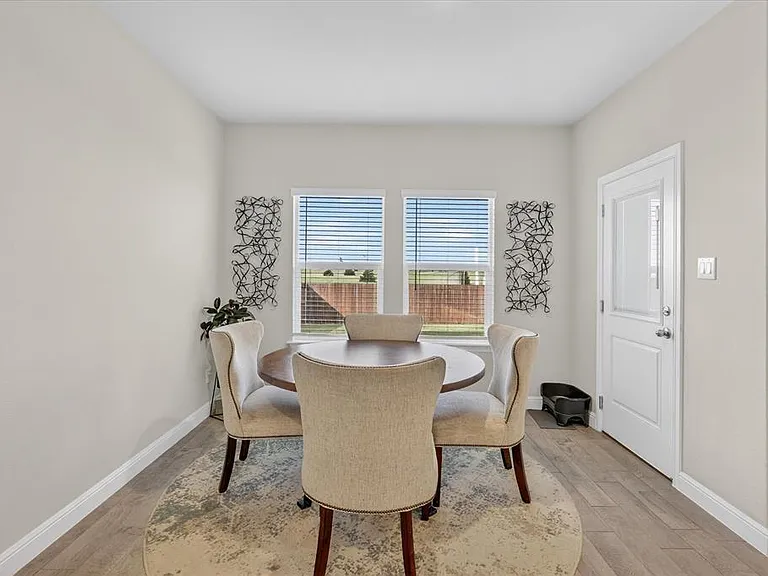 Bright dining area featuring wood-finish flooring, two large windows with horizontal blinds, and a white paneled door with a frosted glass insert
