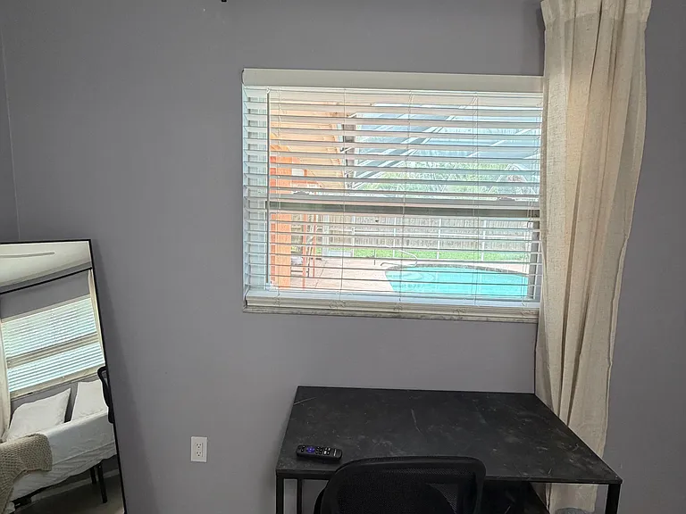 Bedroom workspace area with a desk and chair, plus a window with blinds and curtains overlooking the backyard/pool.