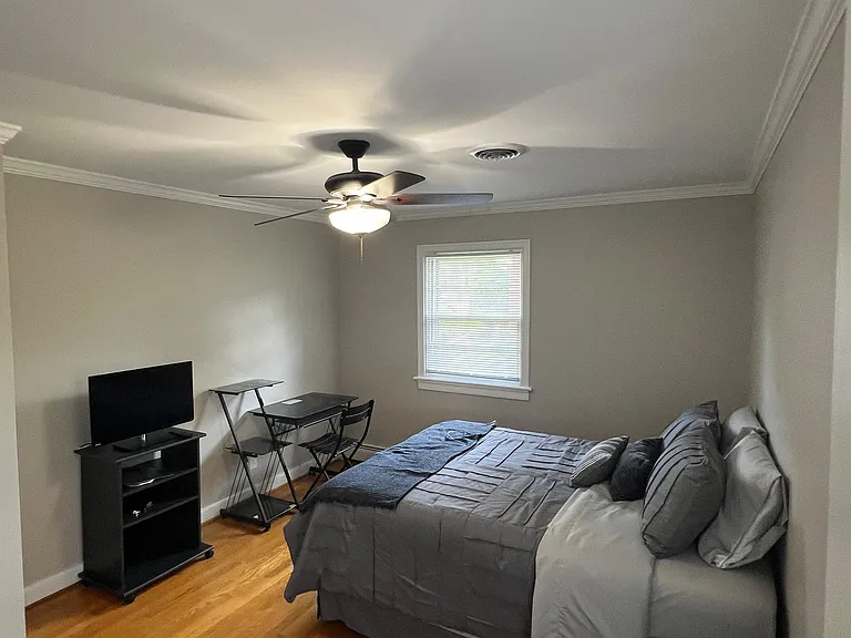 Furnished bedroom with hardwood floors, ceiling fan, and a window with blinds, plus space for a desk and TV.