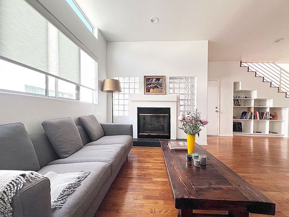 Bright open-concept living space with real wood floors, a working gas fireplace, a skylight, and large windows with roller shades, accented by curated artistic furniture.