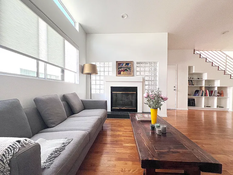 Bright open-concept living space with real wood floors, a working gas fireplace, a skylight, and large windows with roller shades, accented by curated artistic furniture.