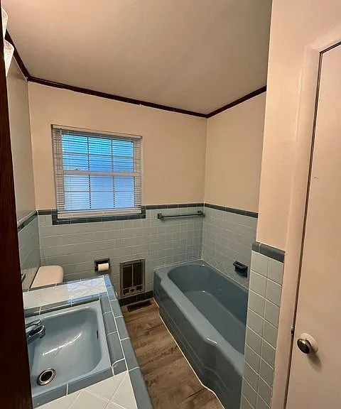 Vintage-inspired bathroom featuring original powder blue ceramic tile matching retro bathtub with coordination built-in since surround. The space offers a clean, well-maintained aesthetic with updated LVP flooring, natural light from large window, and a c