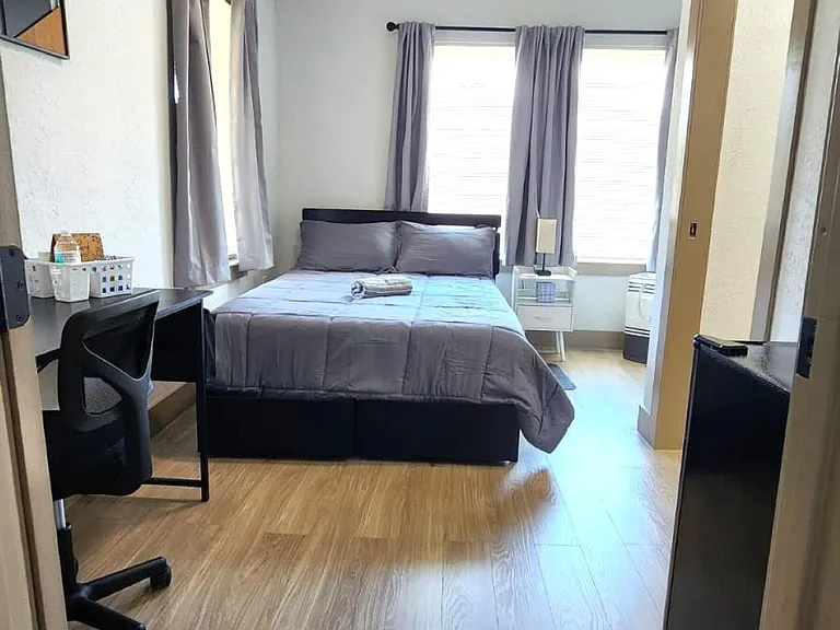 Private bedroom with a full bed, work desk, ceiling fan, and wood-look flooring, plus large windows with curtains.