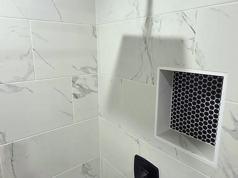Modern shower features elegant white marble-patterned tiles, a black rainfall showerhead, and a convenient built-in niche with stylish hexagonal tiles, offering both functionality and contemporary design.