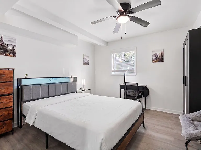 Furnished bedroom with a queen bed, dresser, wardrobe, and a dedicated desk area plus ceiling fan and window blinds.