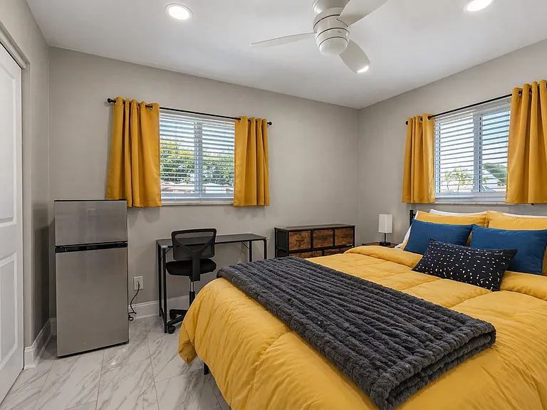 Furnished bedroom with two windows, ceiling fan, and dedicated desk area, plus a mini fridge and dresser for added storage.