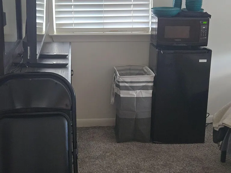 Carpeted bedroom corner with a window and blinds, plus a compact mini-fridge and microwave setup for added convenience.