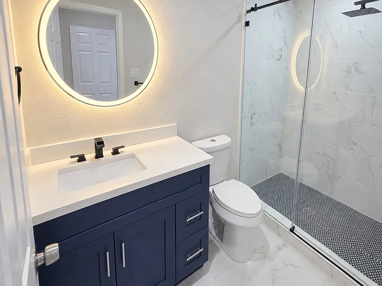 Full bathroom features a sleek vanity with a white countertop, a stylish round backlit mirror, and a spacious glass-enclosed shower with tile and a rainfall showerhead.