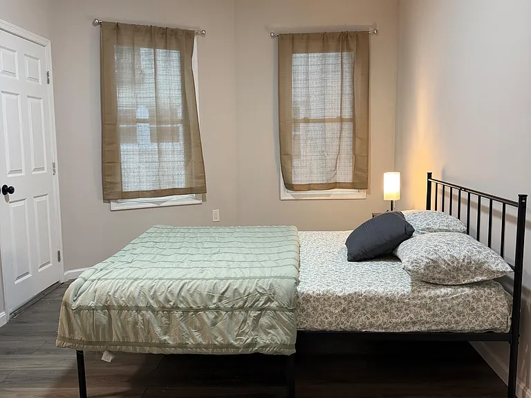 Furnished bedroom with a full-size bed, side table lamp, and two windows with curtains for privacy and natural light.