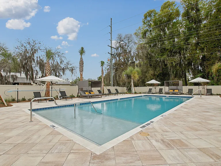 Luxury outdoor pool area with sleek sun loungers, umbrellas, and lush landscaping under a clear blue sky.