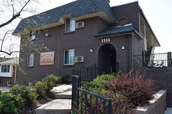 Charming brick apartment building with inviting archway entrance, surrounded by lush greenery on a sunny day.