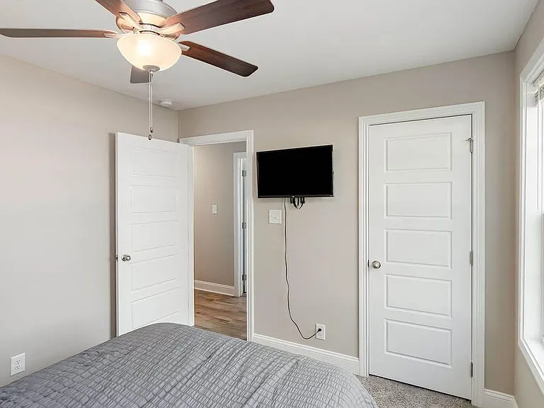 Private bedroom with neutral finishes, ceiling fan, wall-mounted TV, and easy access to the hallway and nearby rooms.