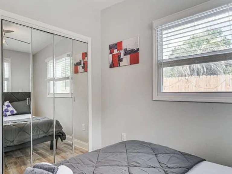 Bedroom with mirrored closet doors, wood-look flooring, and a large window with blinds overlooking the fenced yard.