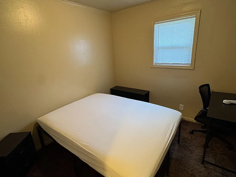 Private bedroom featuring a bed, nightstand, and dedicated desk area, plus a window with blinds for added privacy.