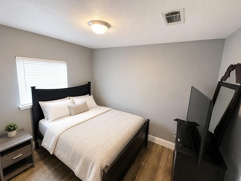 Well-appointed bedroom featuring a queen bed, nightstand, and TV setup, with wood-look flooring and a window with blinds.