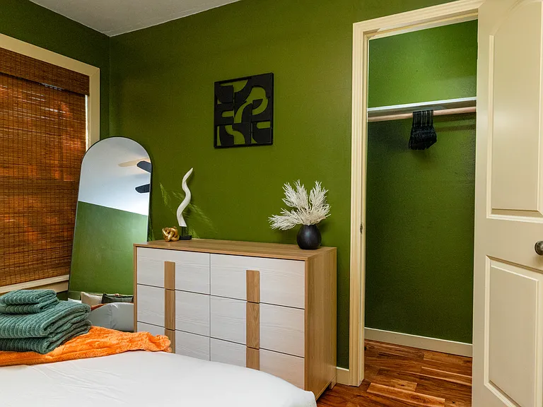 Relax in this serene bedroom featuring rich green walls, warm wood floors, and a plush bed with cozy accent pillows. A ceiling fan, natural textures, and soft lighting create a peaceful, modern retreat.