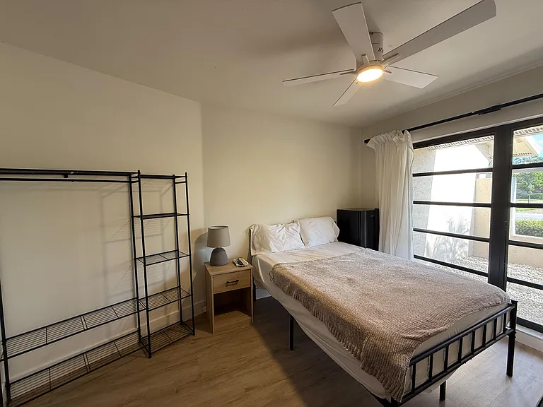 Furnished bedroom with a full-size bed, nightstand, and open shelving, plus a large window and ceiling fan for comfort.