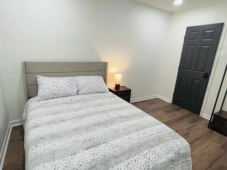 Furnished bedroom with a full bed, nightstand lighting, wood-look floors, and an open clothing rack for easy storage.