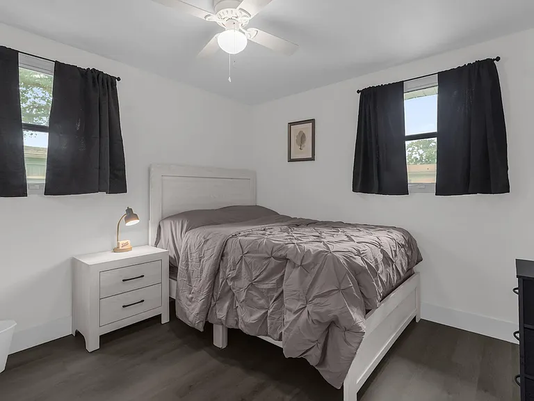 Furnished bedroom with a full bed, nightstand, and ceiling fan, plus two windows with blackout curtains for privacy.