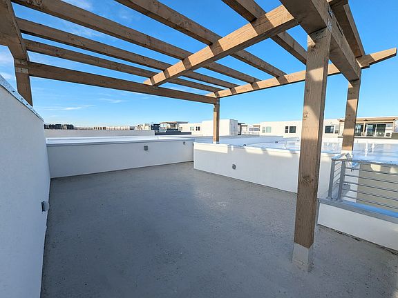 A rooftop terrace with a wooden pergola, white walls, and a concrete floor. The sky is clear and blu