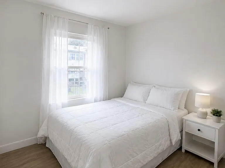 Serene bedroom with a full-size bed, nightstand lighting, and a large window with sheer curtains for natural light.